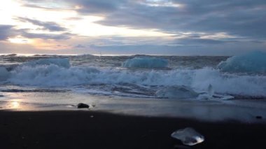 Dalgalar üzerinde rulo buzdağları ve mavi ve beyaz buz parçaları. Dalgalar üzerinde buz kırma. Kumsalda güneş doğarken sörf. İzlanda. Ağır çekim