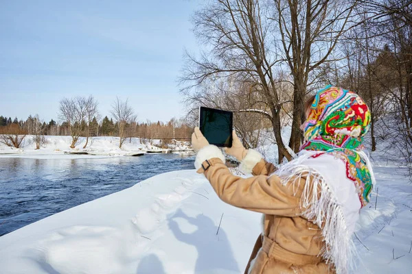 Young beautiful girl in winter outfit is photographing nature on the tablet. Girl looks through the winter photos on the gadget
