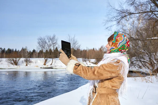 Young beautiful girl in winter outfit is photographing nature on the tablet. Girl looks through the winter photos on the gadget