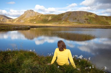 Genç bir kadın bir dağ Gölü kıyısında oturuyor. Bulutlar dağlar ve mavi gökyüzünün suya yansıyan. Seyahat etmek yaz manzarası. İzlanda Europe