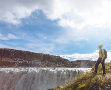 Genç bir kadın İzlanda dev Dettifoss şelaleye bakan bir uçurumun kenarında üzerinde sarı duruyor giymiş. Güneşli yaz tatil günü