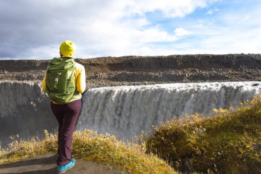 Genç bir kadın İzlanda dev Dettifoss şelaleye bakan bir uçurumun kenarında üzerinde sarı duruyor giymiş. Güneşli yaz tatil günü