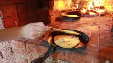 Ulusal elbiseli bir kız şöminede yemek yapıyor. Tavada krepler fırında ateşte kızartılır.