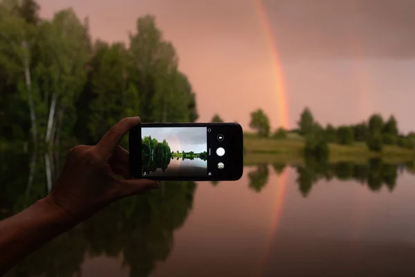 Photographing a landscape on a smartphone. Rainbow and red-yellow sunset with reflection in a forest pond.