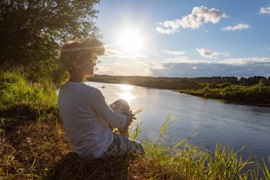        .  Kafasında çelenk olan bir kız nehir kenarında oturur. Çerçevede güneş ışığı       .      