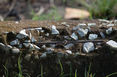 Kırık güçlendirilmiş beton kirişe yakın çekim. Toplam taşları ve paslı demirleri gösteriyor. Etrafında yeşil çimenler büyüyor..