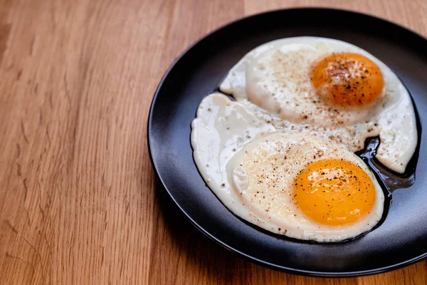 two fried egg with black pepper on black plate on wooden table copy space for text