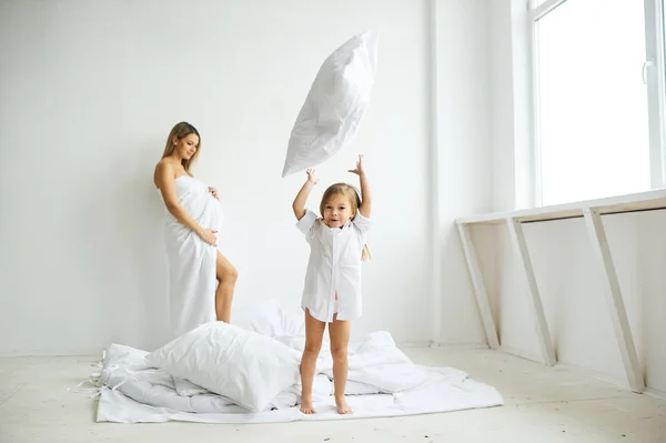 Uma bela mãe grávida e filha brincando em uma cama branca em — Fotografia de Stock