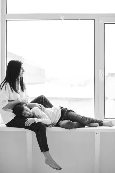 Young beautiful mother gently hugs her son on the windowsill — Stock Photo, Image