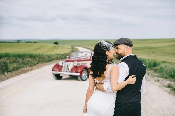 Elegante pareja de boda, novia, novio caminando en un campo nea carretera —  Fotos de Stock