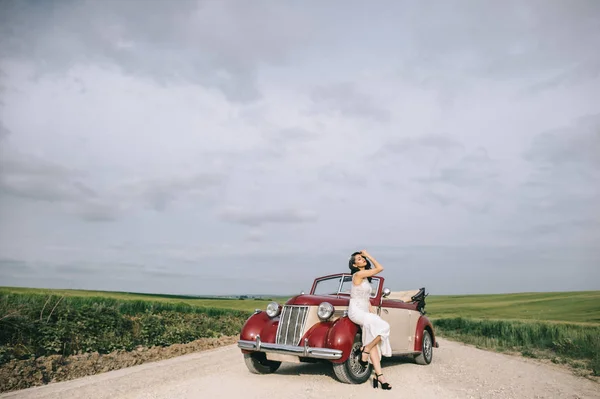 Stijlvolle mooie bruid zittend op een rode retro auto op een veldweg. — Stockfoto