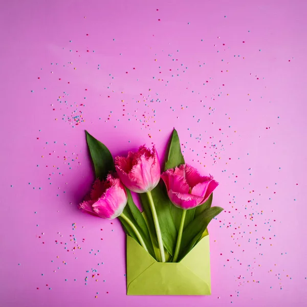 Drei rosa Tulpen in hellgrünem Umschlag auf rosa Hintergrund, verziert mit süßem Konfetti. — Stockfoto