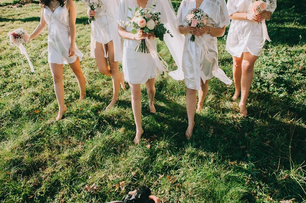 Faceless bridesmaids and bride in satin robes with bouquets in hands walk barefoot on the grass. — Stock Photo, Image