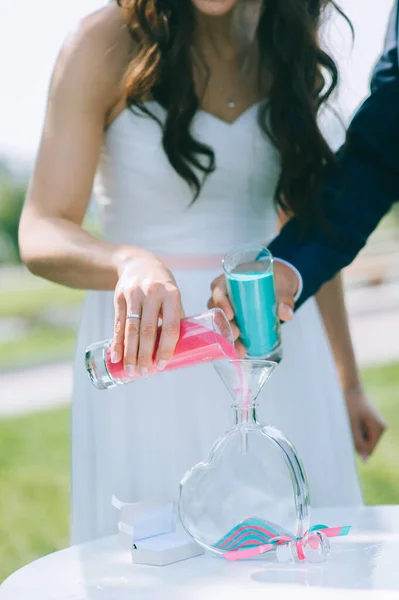 Primer plano de la novia y el novio vertiendo arena de colores en un jarrón de vidrio en forma de corazón. —  Fotos de Stock