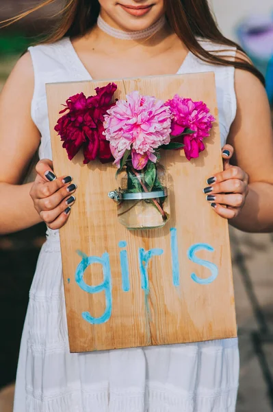 Niña sosteniendo una tabla de madera con la inscripción girla y adjunta a ella un frasco de vidrio con peonías. — Foto de Stock