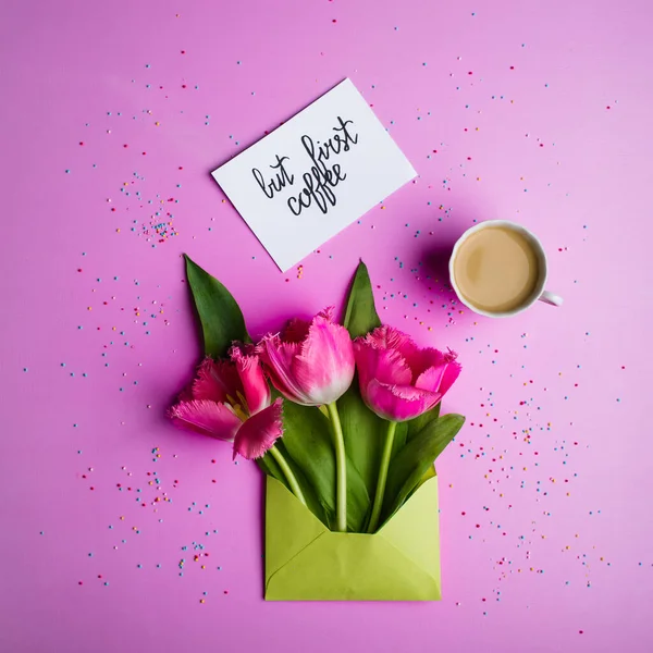 Tulips in a light green envelope, a cup of coffee with milk and a card with the inscription. — Stock Photo, Image