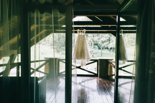 Вид через открытое окно на шикарное оригинальное свадебное платье, висящее на вешалке на террасе. — стоковое фото