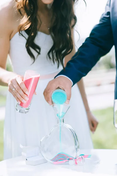 Primer plano de la novia y el novio vertiendo arena de colores en un jarrón de vidrio en forma de corazón. —  Fotos de Stock