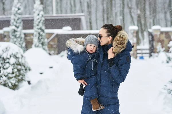 A portret of beautiful young mother dressed in blue jacket with a child in her arms in winter near the house with a snow covered christmas trees in a background — Stock Photo, Image