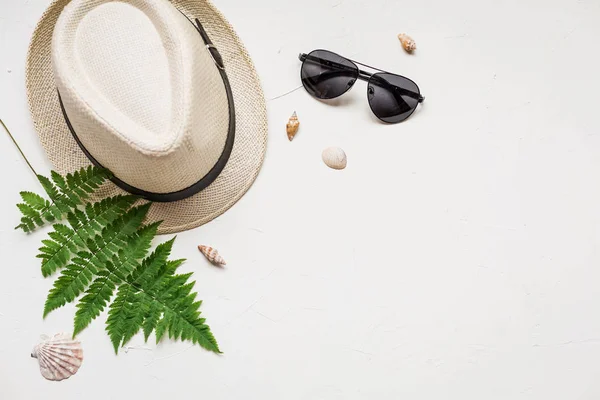 Summer flat lay with a hat and a green fern, a red sea star, seashells and glasses on white concrete background.