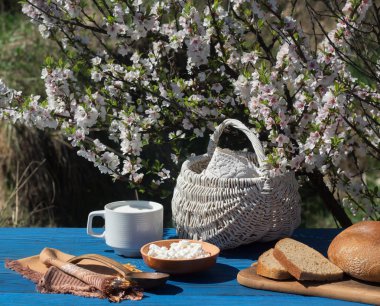 Sepet, süt, peynir ve ekmek bir çiçekli çalı arka planı mavi kurullarının bir masada bir fincan