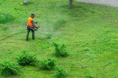Turuncu yelek giyen bir kamu görevlisi el gazı makinesiyle parkta çim biçiyor.