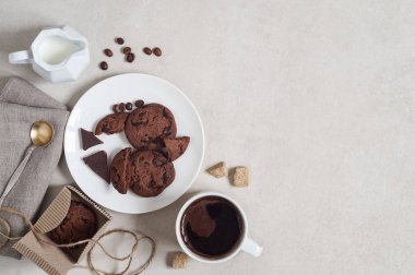 Beyaz tabakta kakao ve çikolatalı yulaflı kurabiyeler ve karton bir kutuda, bir fincan kahve ve bir sürahi süt.