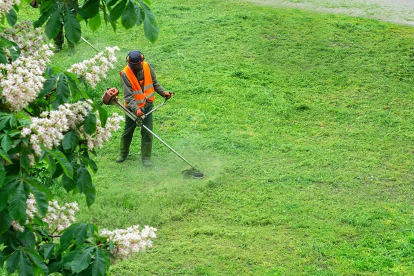 Turuncu yelek giyen bir kamu görevlisi el gazı makinesiyle parkta çim biçiyor.