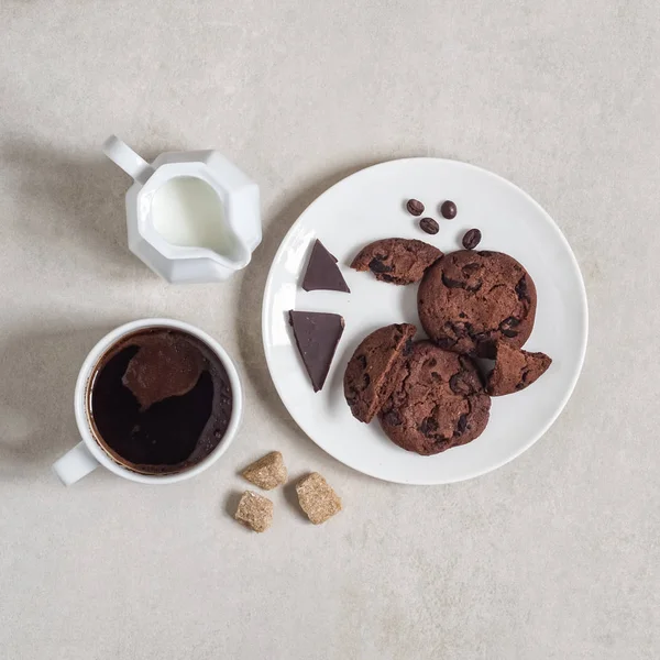 Beyaz tabakta kakaolu ve çikolatalı yulaflı kurabiyeler, bir fincan kahve ve süt sürahisi.