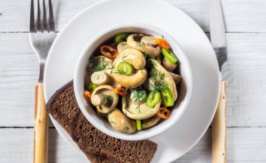 Beyaz ahşap bir masada biber ve otlarla turşulu ya da tuzlu beyaz mantarlar.