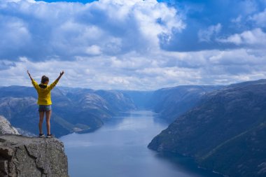 Kolları kaldırıp sarı ceketli kız Preikestolen, Norveç'ten Lysefjord fiyortuna bakarak