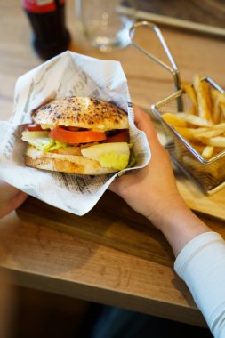 Kağıda sarılmış ızgara tavuk burger, görünür domates, marul ve turşu tutan birinin yakın çekimi. Bir porsiyon altın kızartma yakınlardaki metal bir sepette servis edilir..