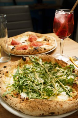 Bir İtalyan restoranında servis edilen taze roka ve krema soslu ahşap kaplı pizzanın yakın çekimi. Arka planda prosciutto ve kırmızı Aperol Spritz kokteyli olan bir pizza daha..