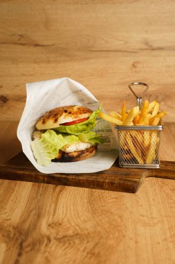Kızarmış tavuklu sandviç, kıvırcık salata ve domatesli. Yanında da metal bir sepetin içinde patates kızartması var. Hepsi de ahşap bir tahtanın üzerine..