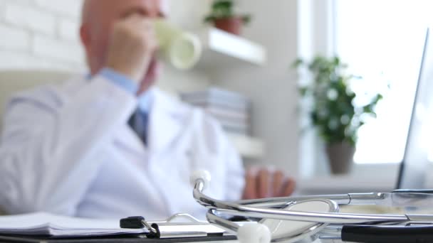 深刻な医師は聴診器フォーカスで机の上で不鮮明な画像でコーヒーを飲む — ストック動画