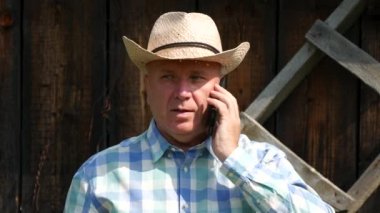 Çiftçi arka planda ahşap bir ahır ile cep telefonuna el hareketi ciddi konuşmak