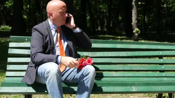 坐在长凳上的男子等待着玫瑰在他手中的日期 并与移动交谈 — 图库视频影像