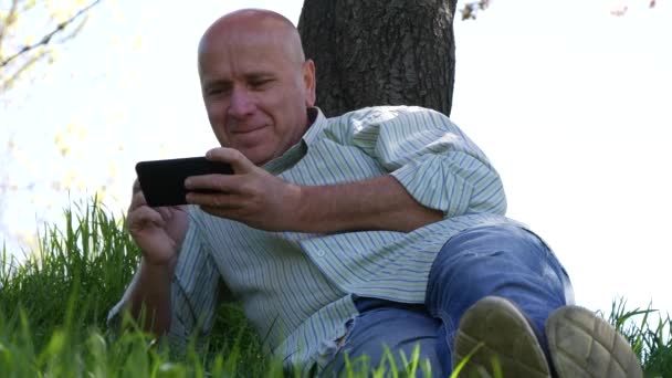 Расслабленный Человек Лежит Зеленой Траве Использовать Мобильный Сенсорный Экран Улыбаясь — стоковое видео