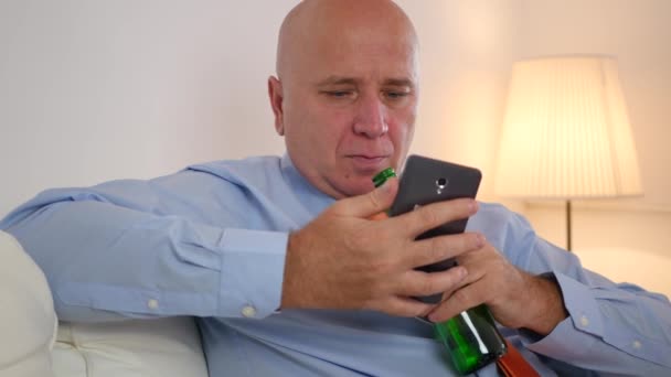男人坐在沙发上看手机新闻和喝啤酒 — 图库视频影像