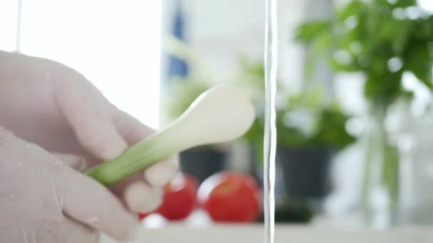 Movimento lento com o homem na cozinha vestindo luvas em suas mãos lavando cebola verde com água limpa — Vídeo de Stock