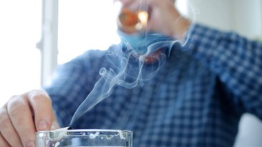 Yüzüne koruyucu maske takmış bir adam bir barda alkollü bir bardak içer ve sigara içer.