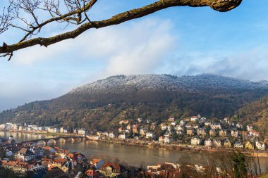 Buz kaplı tepeler, geleneksel evler ve Neckar Nehri üzerindeki Eski Köprü ile Heidelberg manzaralı açık bir kış akşamı.