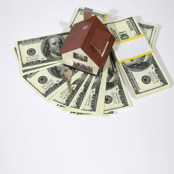 A small house lies on a fan of hundred dollar bills. The keys to the purchased house. Reduced copy of the house on a white background.