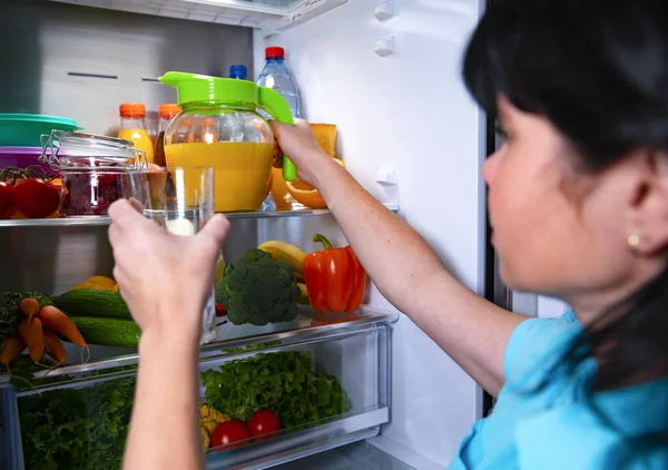 Open fridge. A woman holds a glass in her hand and pours fresh juice from a jug. Fresh vegetables and fruits.
