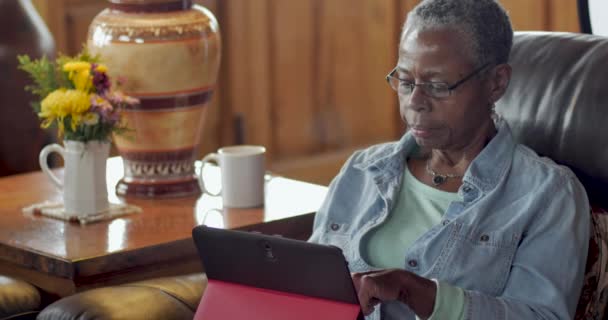 Пожилая пожилая черная женщина использует свой цифровой планшет в своей гостиной — стоковое видео