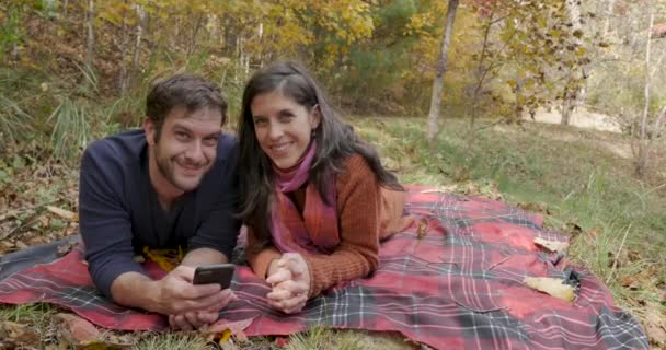 Pareja mirando a la cámara y sonriendo tumbada sobre una manta en un parque — Vídeos de Stock