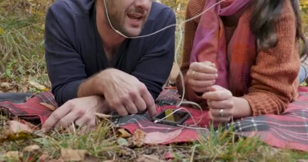 Pareja compartiendo la misma música mientras están acostados en una manta afuera — Vídeo de stock