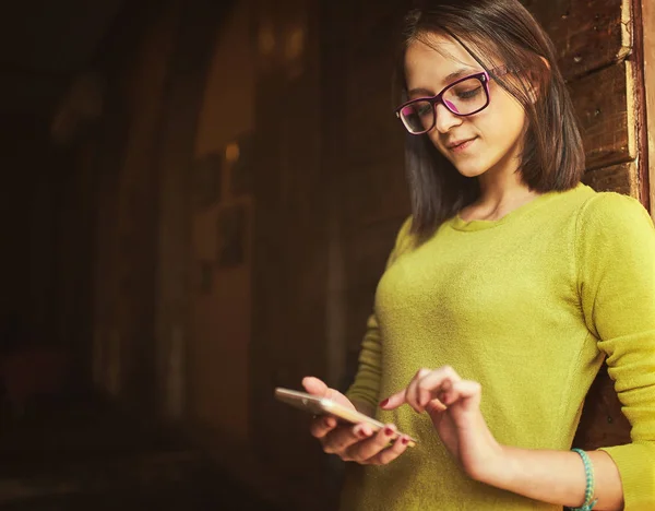 Cheerful young female student with cute smile siting in modern cafe interior using cell phone