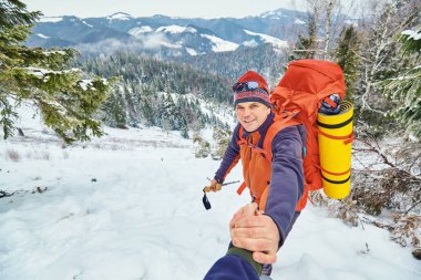 Young tourists with backpacks, athletic boy helps slim girl to clime rocky mountain top against bright summer sky and mountain range background.
