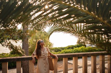 İnce esmer, açık tenli ve sandaletli. Balyalara yaslanmış ve büyük palmiye yaprağının altına bakıyor. Hırvatistan 'da yaz tatilinde Brac Adası' nı gezen elinde saman çantasıyla zinde bir kadın..